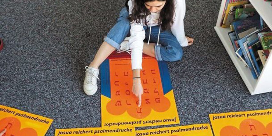 Ein Mädchen sitzt im Schneidersitz und zeigt auf ein hebräisches Poster, umgeben von Büchern.