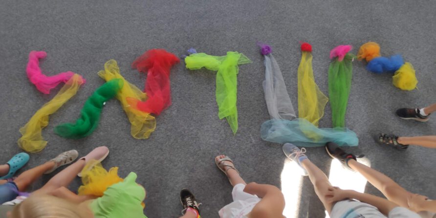 Kinder stehen im Kreis um bunte Tücher, die auf grauem Teppich das Wort UNITY bilden.