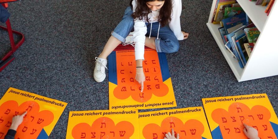 Eine Frau sitzt mit Kindern am Boden. Sie zeigen auf bunte Poster mit hebräischen Buchstaben, Bücher im Regal.