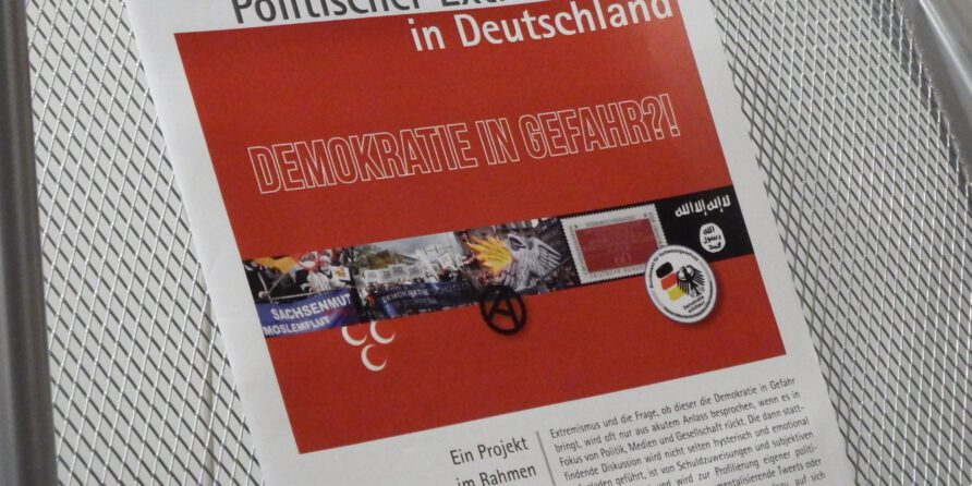 Broschüre „Politischer Extremismus in Deutschland“ mit Flaggen liegt auf einem Metallstuhl.