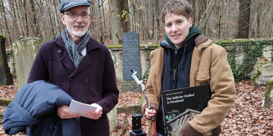 Zwei Personen im herbstlichen Friedhof; sie halten Stift, Papier, Buch und Kamerastativ. Grabsteine im Hintergrund.