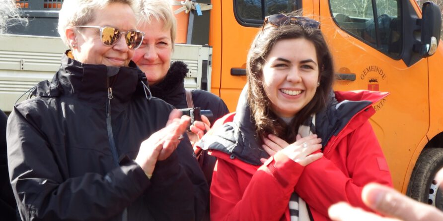 Drei Frauen vor einem orangefarbenen LKW, lächelnd und klatschend; eine trägt Rot und hält sich an die Brust.