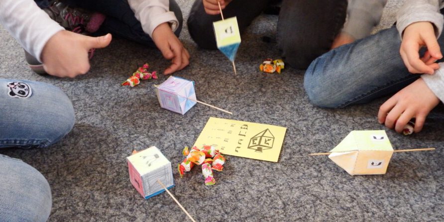 Drei Kinder spielen auf grauem Teppich mit bunten Dreideln und Süßigkeiten; eins zeigt Daumen hoch.