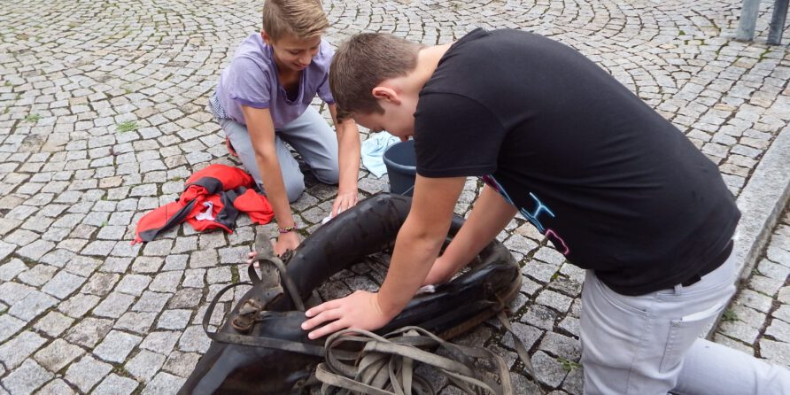 Zwei Jungen knien auf Kopfsteinpflaster und helfen einer großen, dunklen Robbenfigur mit Gurten.