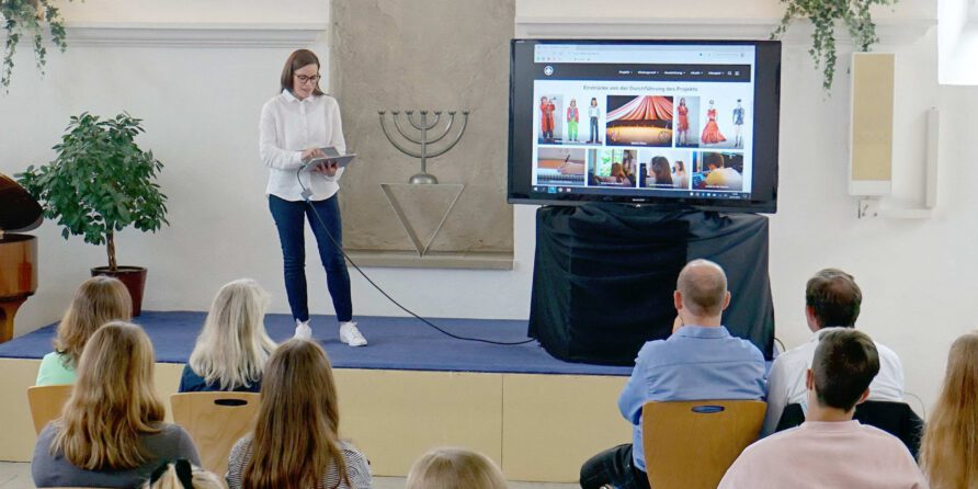 Eine Frau präsentiert mit Tablet auf einer Bühne; bunte Bilder, Pflanzen und Menora sind zu sehen.