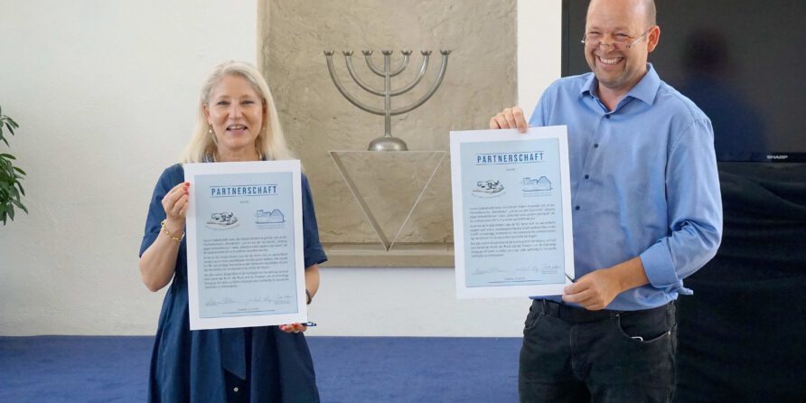 Eine lächelnde Frau und ein Mann halten Zertifikate vor einer großen Menora und Steinwand.