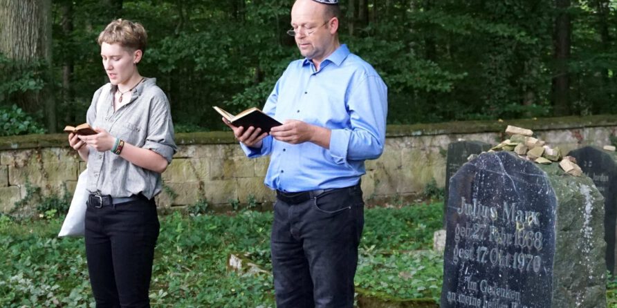 Zwei Personen lesen an einem alten Grabstein mit kleinen Steinen in einem ruhigen, grünen Friedhof.
