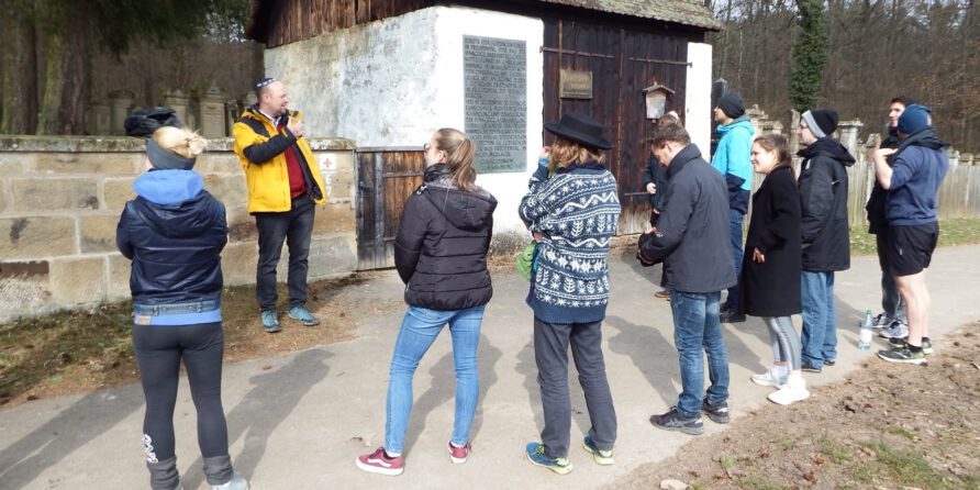 Eine Gruppe lauscht einem Führer vor einem alten Holzhaus mit Schild, umgeben von Bäumen und Mauern.