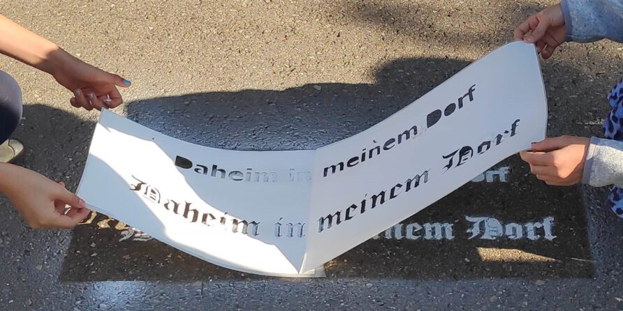 Zwei Personen halten eine Schablone mit „Daheim in meinem Dorf“, deren Schatten auf dem Boden erscheint.