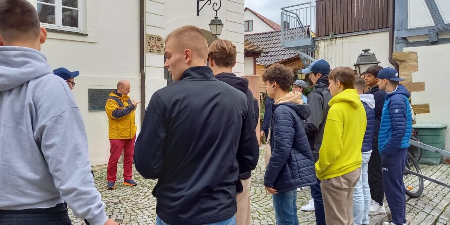 Eine Gruppe junger Männer hört draußen auf Kopfsteinpflaster einem Mann im gelben Jackett zu.