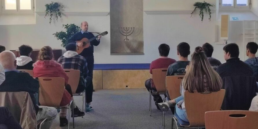 Ein Mann spielt Gitarre und singt vor sitzendem Publikum; hinter ihm hängt ein Menora an der Wand.