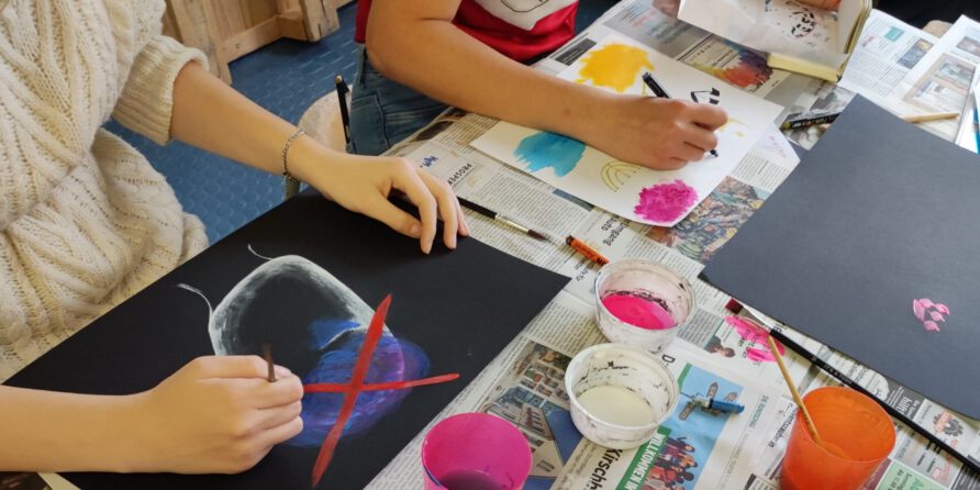 Zwei Personen malen auf schwarzem Papier am Zeitungs-Tisch, Pinsel und Farben liegen überall.