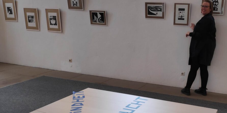 Eine lächelnde Person hängt gerahmte Schwarzweißkunst auf; am Boden steht KINDHEIT und FLUCHT.