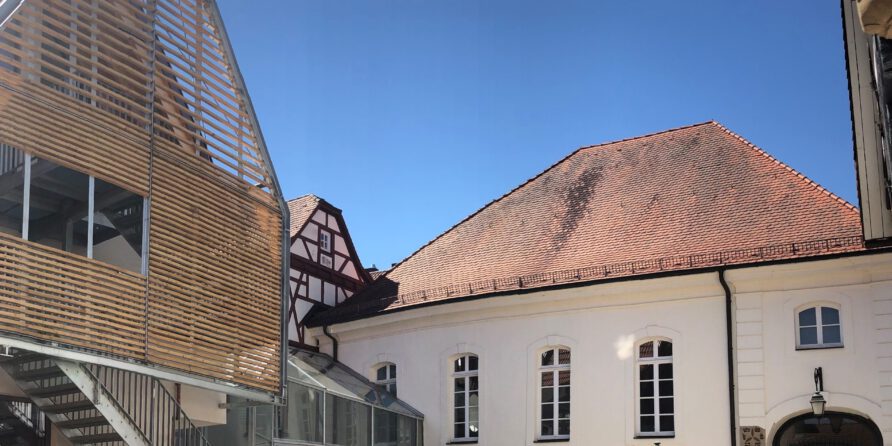 Sonniger Hof mit modernem Gebäude und Holzpaneelen links, klassischem Haus mit roten Ziegeln rechts.