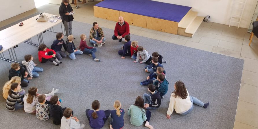 Kinder sitzen im Kreis auf Teppichboden, zwei Erwachsene daneben, wohl in einem Klassenraum.
