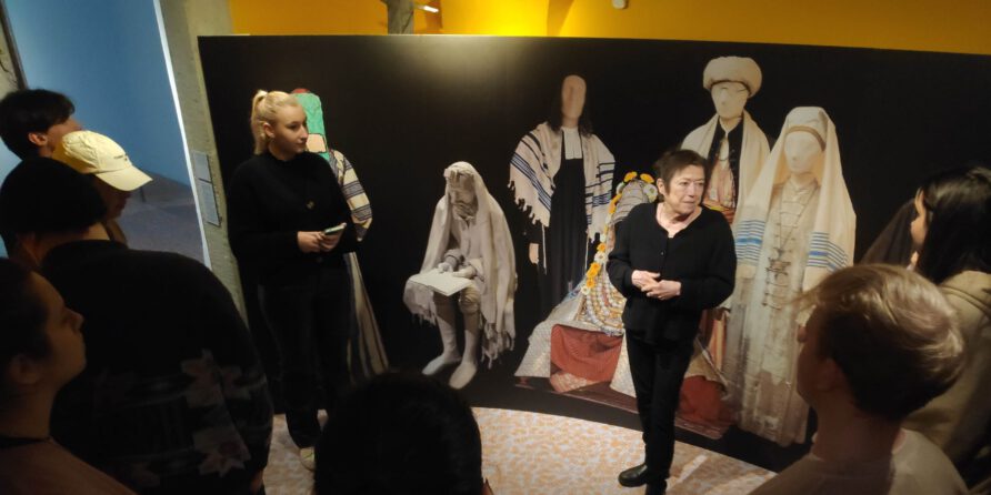 Eine Gruppe hört einer Frau vor Trachtenpuppen im Museum mit schwarzem Hintergrund und gelben Wänden zu.