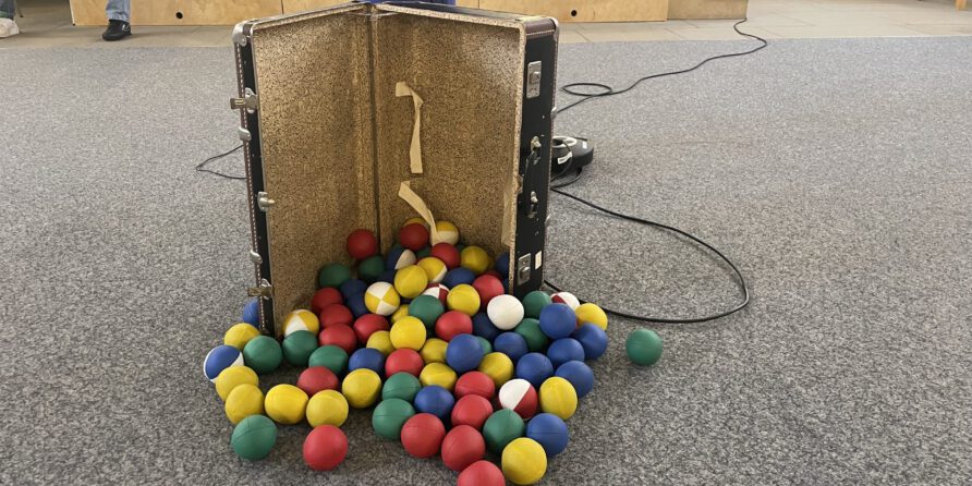 Ein offener Koffer mit bunten Plastikbällen liegt auf Teppich, hinten sind schwarze Kabel zu sehen.