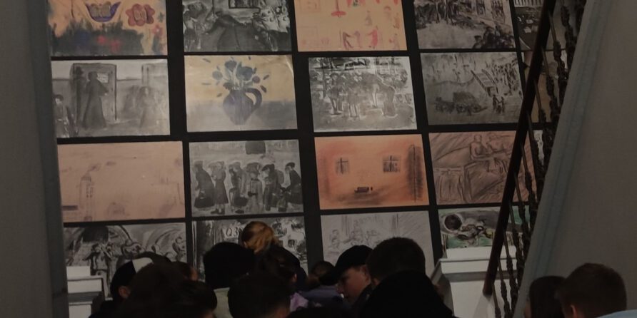 Eine Gruppe steht vor einer Wand mit mehreren Reihen Schwarz-Weiß- und Sepiazeichnungen, vom Treppenhaus aus gesehen.