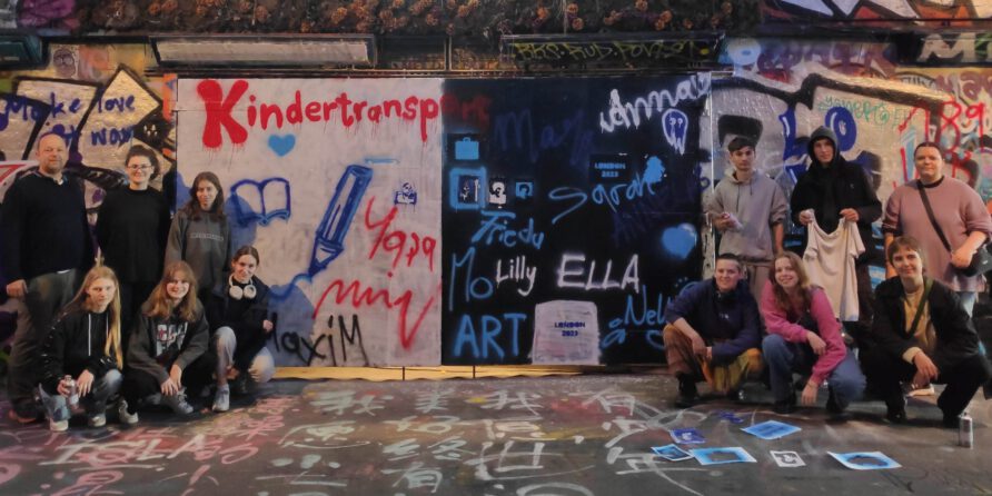 Eine Gruppe posiert vor einer bunten Graffitiwand mit Kindertransp. und Namen, alle zur Kamera.