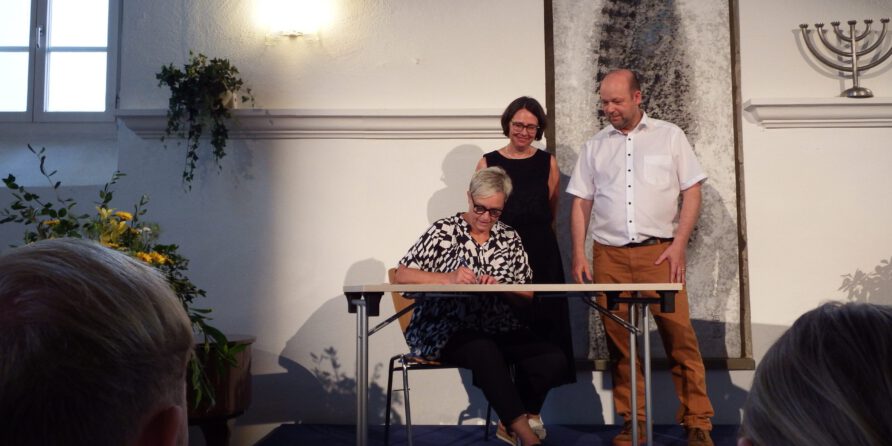 Eine Frau unterschreibt am Schreibtisch, zwei Personen stehen dahinter; heller Raum mit Deko.