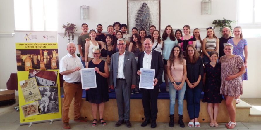 Etwa 25 Personen posieren lächelnd mit Urkunden vor einer Bühne und Banner zu 1700 Jahren Jüdisches Leben.