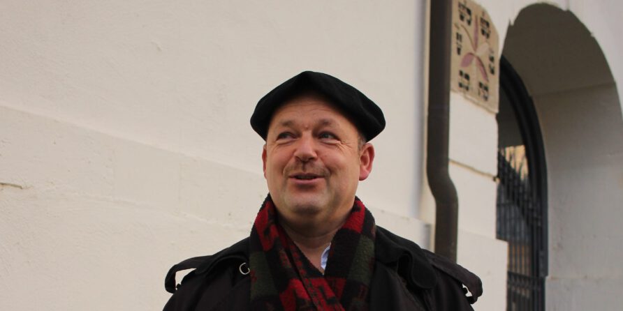 Ein Mann mit schwarzem Barett, dunklem Mantel und Schal lächelt vor heller Wand mit Türbogen.