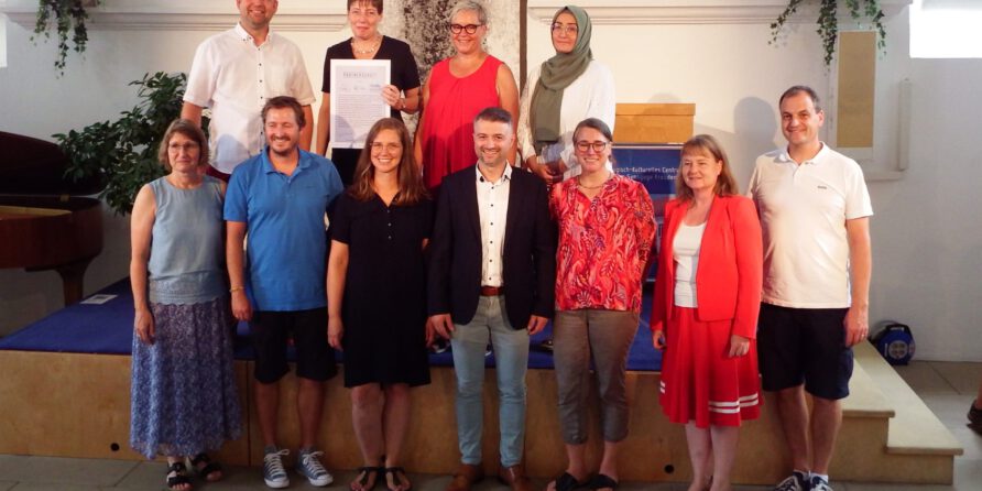 Zwölf Erwachsene lachen für ein Gruppenfoto drinnen, teils auf Podest, eine hält eine Urkunde.