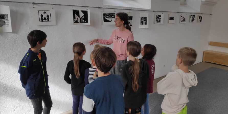 Eine Frau zeigt Kindern schwarz-weiße Kunstwerke an der Wand, die aufmerksam zuhören.