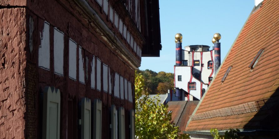 Fachwerkhäuser umrahmen ein modernes Gebäude mit runden goldenen Kuppeln, Bäume und blauer Himmel dahinter.