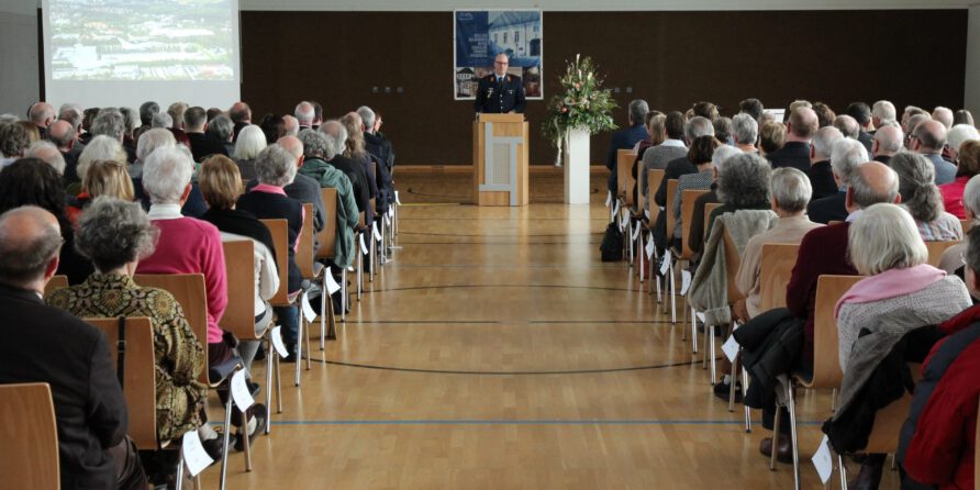Eine große Zuhörerschaft blickt im Saal auf einen Redner am Podium, daneben eine Leinwand und Blumen.