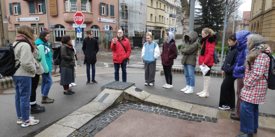 Eine Gruppe steht im Kreis auf dem Bürgersteig und hört einer Person im roten Jacke zu, Stadtumgebung.