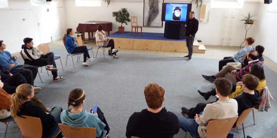 Mehrere Personen sitzen im Kreis, hören einem Sprecher zu; Workshop in hellem Raum mit Fenstern.