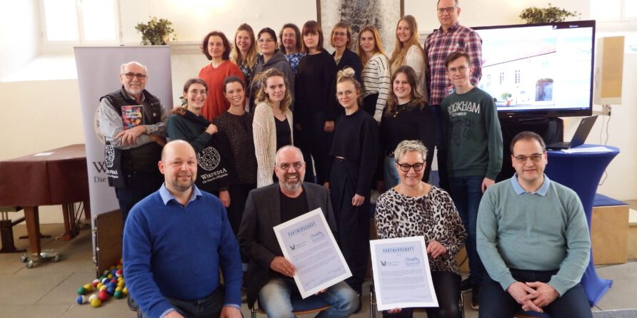20 Personen posieren drinnen, zwei vorne halten Urkunden. Im Hintergrund Banner und Bildschirm.
