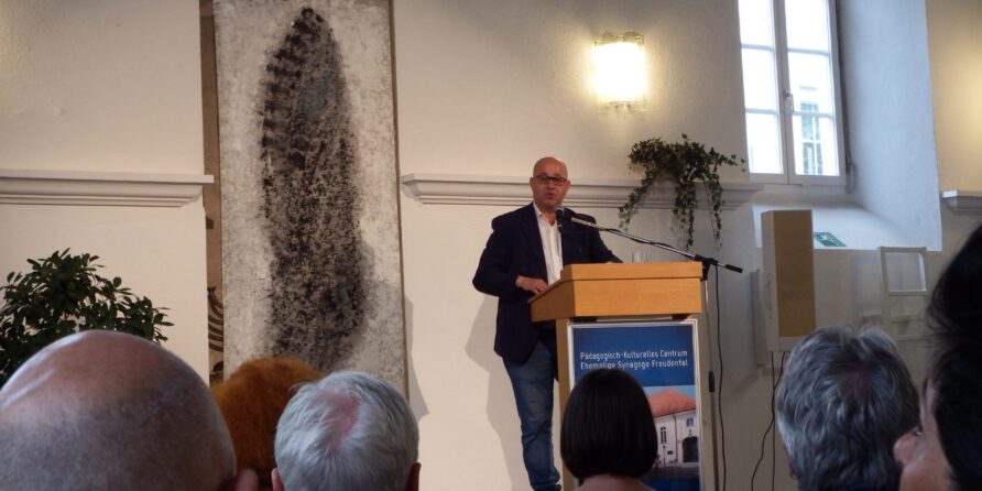 Ein Mann im Anzug spricht am Rednerpult vor Publikum in hellem Raum mit Kunst, Pflanzen und Fenster.