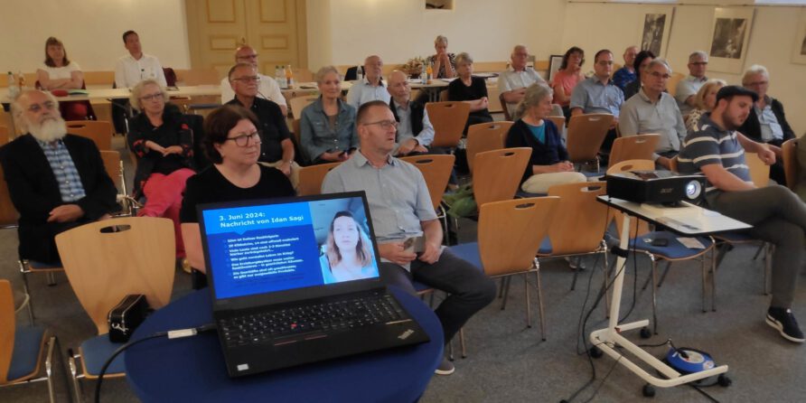 Menschen im Konferenzraum hören einem Vortrag zu, vorne ein Laptop mit Foto und Text einer Frau.