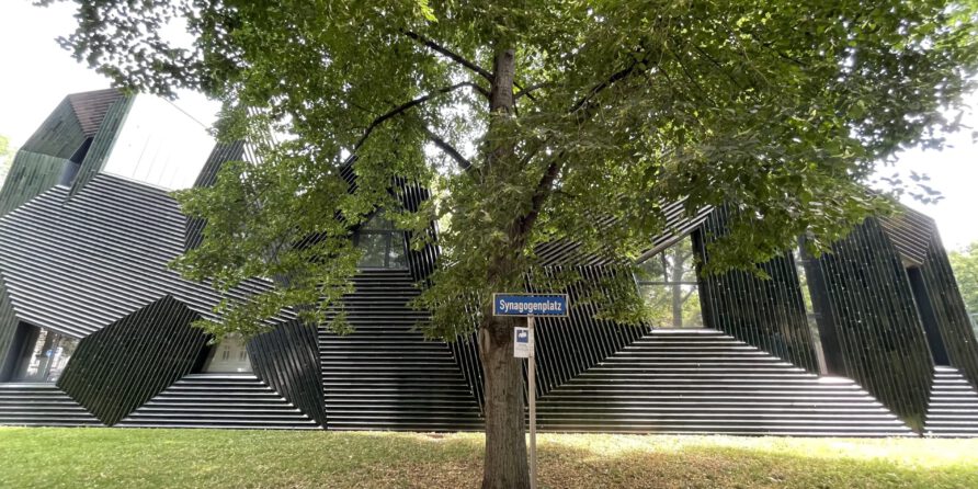 Vor einem modernen, schwarz gestreiften Gebäude steht ein großer Baum; Schild „Synagogenplatz“.