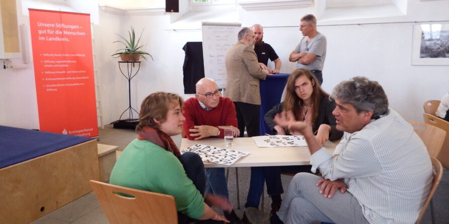 Fünf Personen diskutieren am Tisch mit Unterlagen, zwei stehen am Flipchart. Banner und Fenster im Raum.