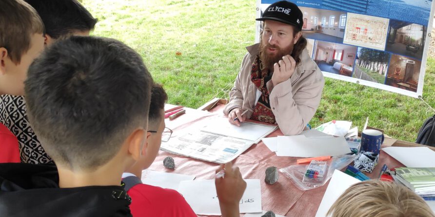 Ein bärtiger Mann mit schwarzem Hut spricht draußen am Tisch mit Kindern, Bastelmaterial liegt bereit.