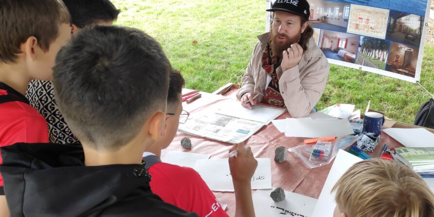 Ein bärtiger Mann mit Mütze spricht am Tisch im Freien mit Kindern, Poster und Gras im Hintergrund.