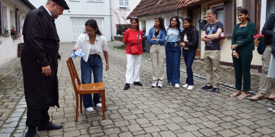 Mehrere Personen stehen im Hof und sehen zu, wie eine Frau einen Stuhl putzt, ein Mann beobachtet sie.
