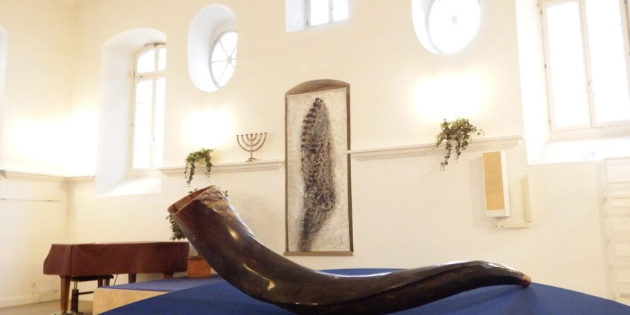 Ein großer Schofar liegt auf blauem Tuch in hellem Raum mit Bogenfenstern, Klavier und Menora.