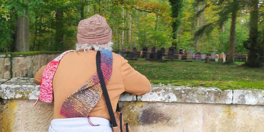 Eine ältere Person im braunen Mantel blickt im stillen, grünen Friedhof über eine moosige Mauer.