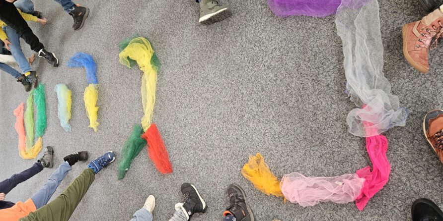 Mehrere Personen im Kreis auf grauem Teppich, bunte Tücher am Boden; nur Beine und Schuhe sichtbar.