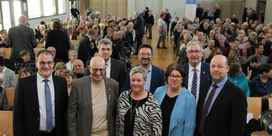 Acht Erwachsene lächeln vorne in einem vollen Saal bei einer formellen Veranstaltung.