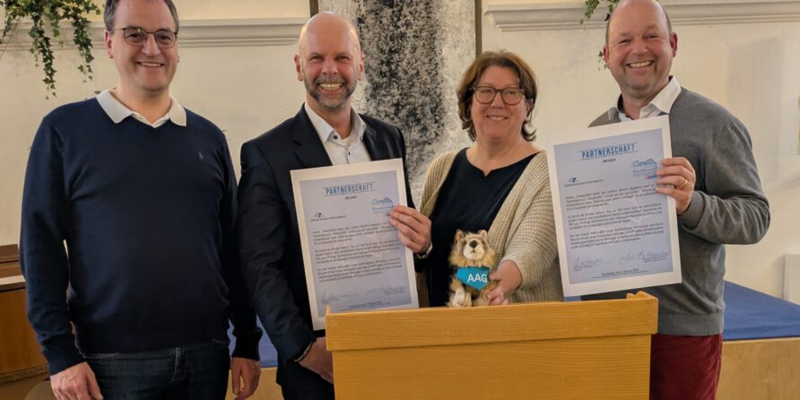 Vier lachende Erwachsene hinter einem Podium, zwei halten Dokumente. Ein Stoffhund und Grün im Hintergrund.