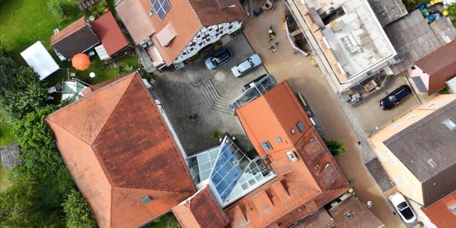 Luftbild: Rote Dächer um Hof mit Glasdach, Autos & Menschen; Solarpanels auf Dächern, Grün drumherum.