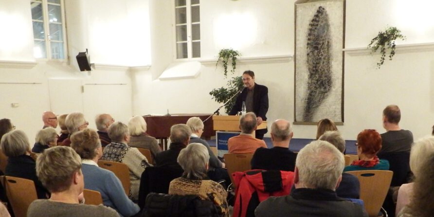 Ein Mann spricht am Rednerpult vor Publikum in einem hellen Raum mit Pflanzen und Kunstwerken.