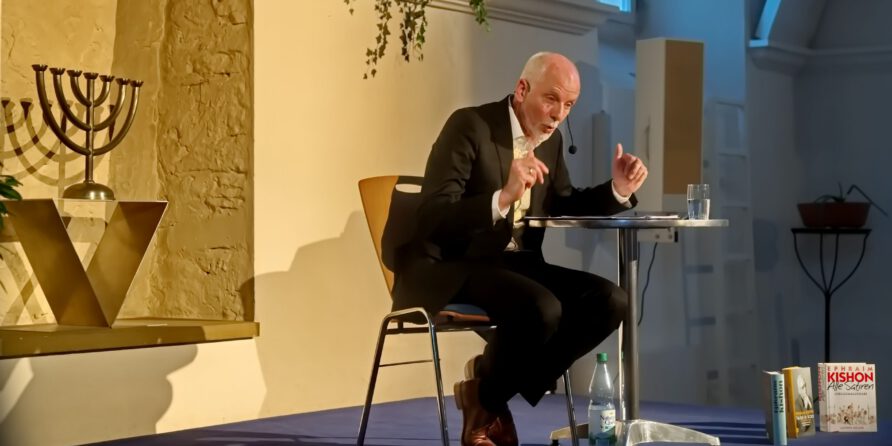 Ein Mann im Anzug spricht mit Headset am Tisch auf der Bühne, Bücher und Menora im Hintergrund.