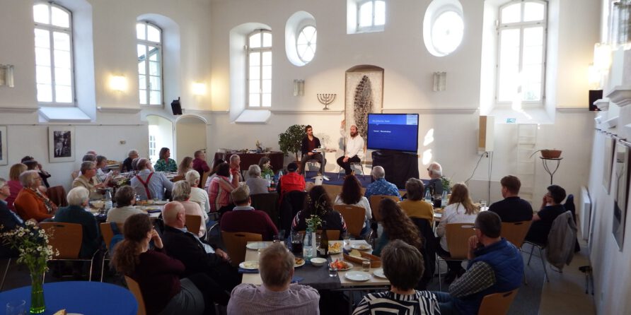 Menschen sitzen an Tischen mit Essen, hören zwei Rednern zu; Präsentation läuft, Menora an der Wand.