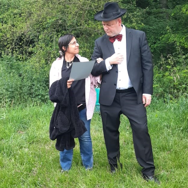 Ein Mann im Anzug und eine Frau mit Papier stehen sich draußen vor grünem Laub gegenüber.